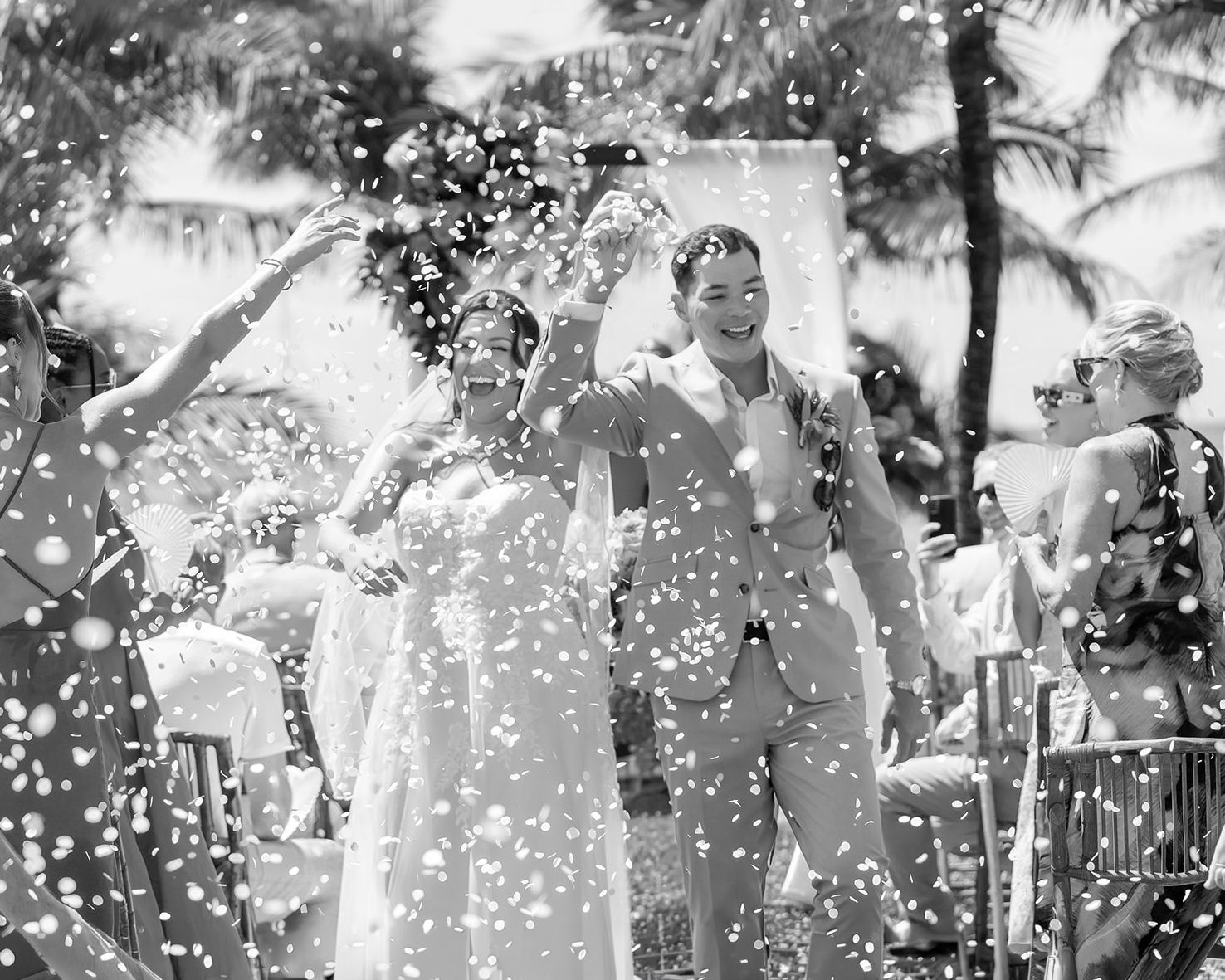 Black and white wedding photo of couple celebrating with confetti at tropical outdoor ceremony.