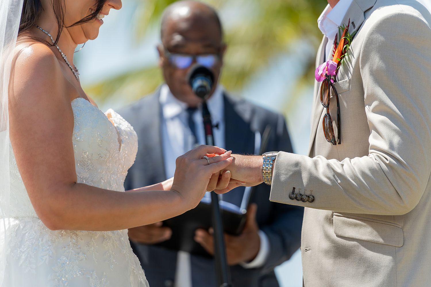 Bride placing wedding ring on grooms finger during outdoor ceremony with officiant in background.