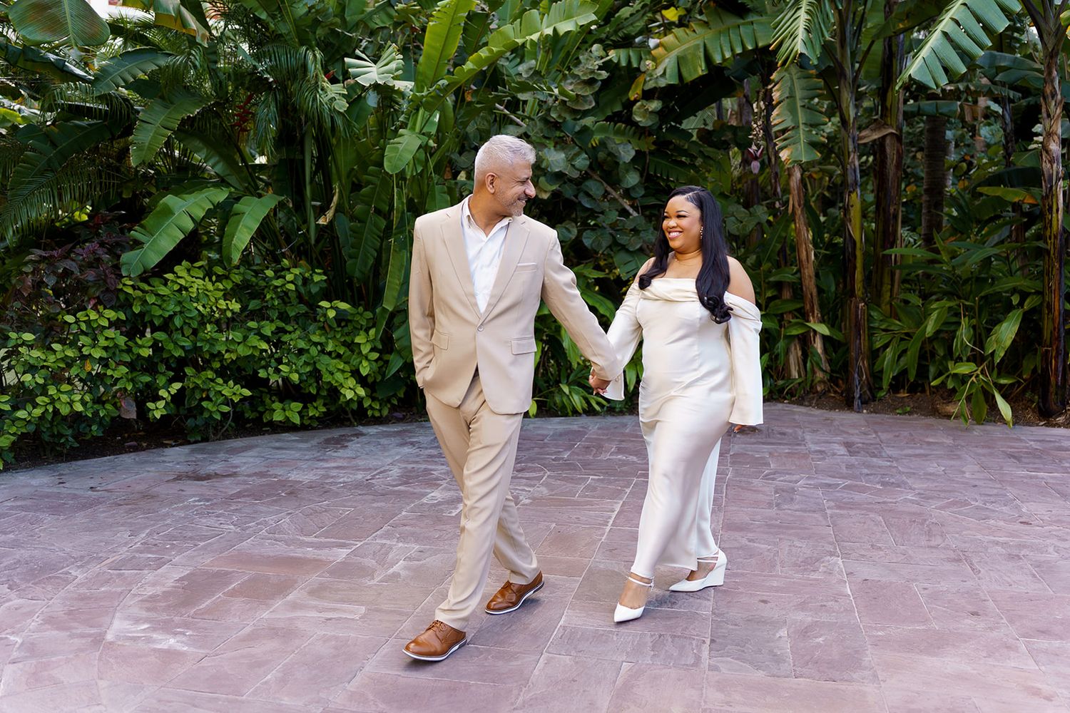 Couple in elegant white and beige outfits holding hands while walking on a patio surrounded by lush tropical greenery.