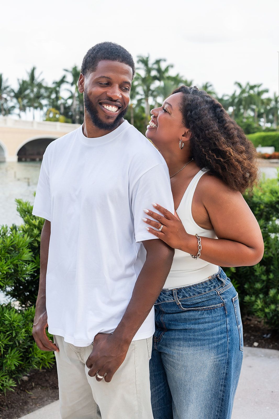 Happy Black couple laughing together outdoors near tropical waterway, woman in white top and jeans embracing smiling man in white tee.