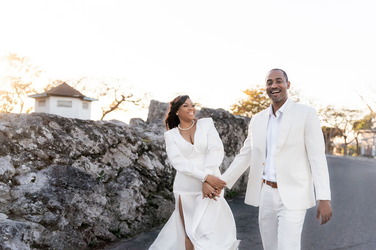 Couple in white attire holding hands and smiling outdoors near stone wall at sunset.