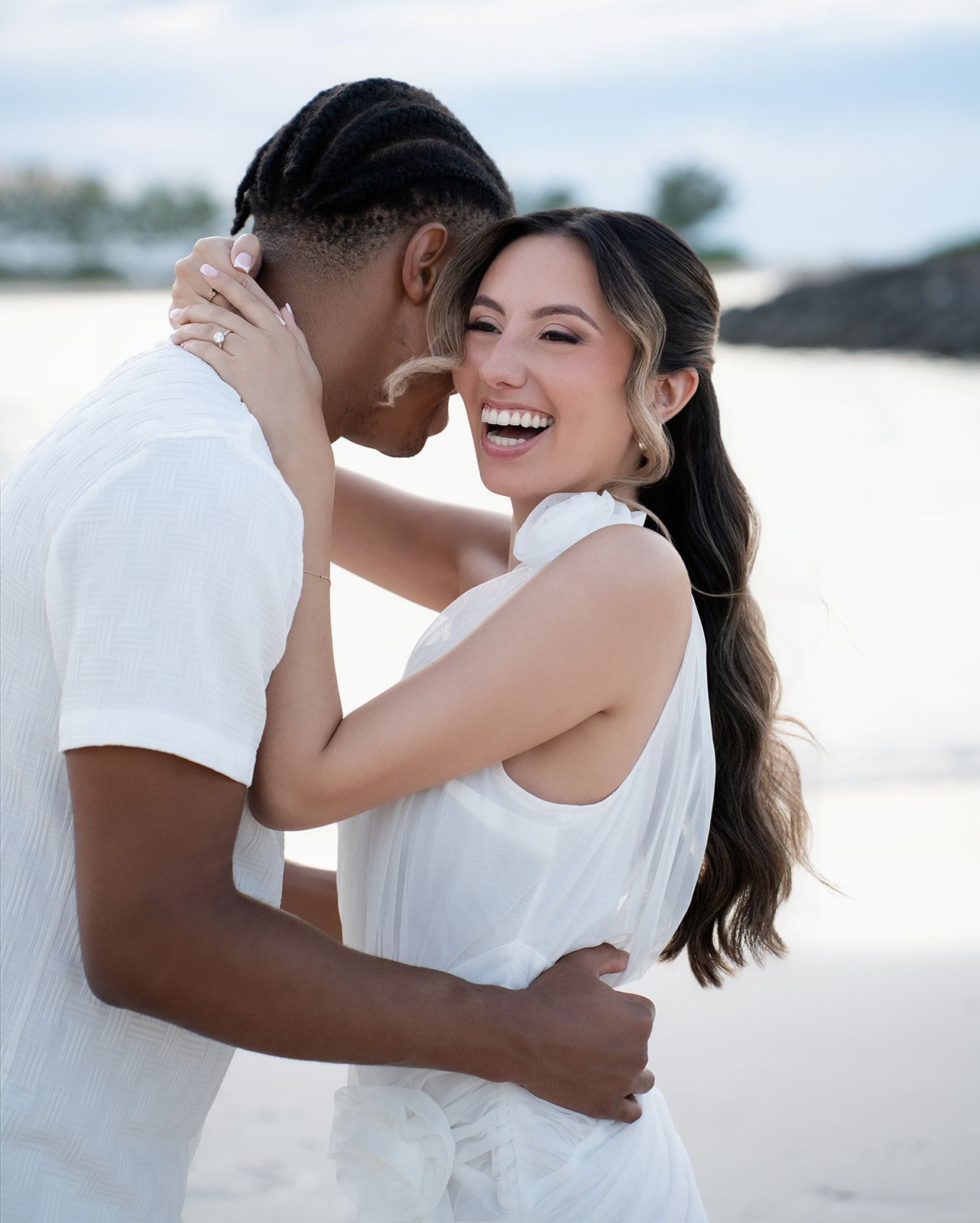 Happy couple embracing and laughing on a beach, woman wearing white dress and engagement ring.