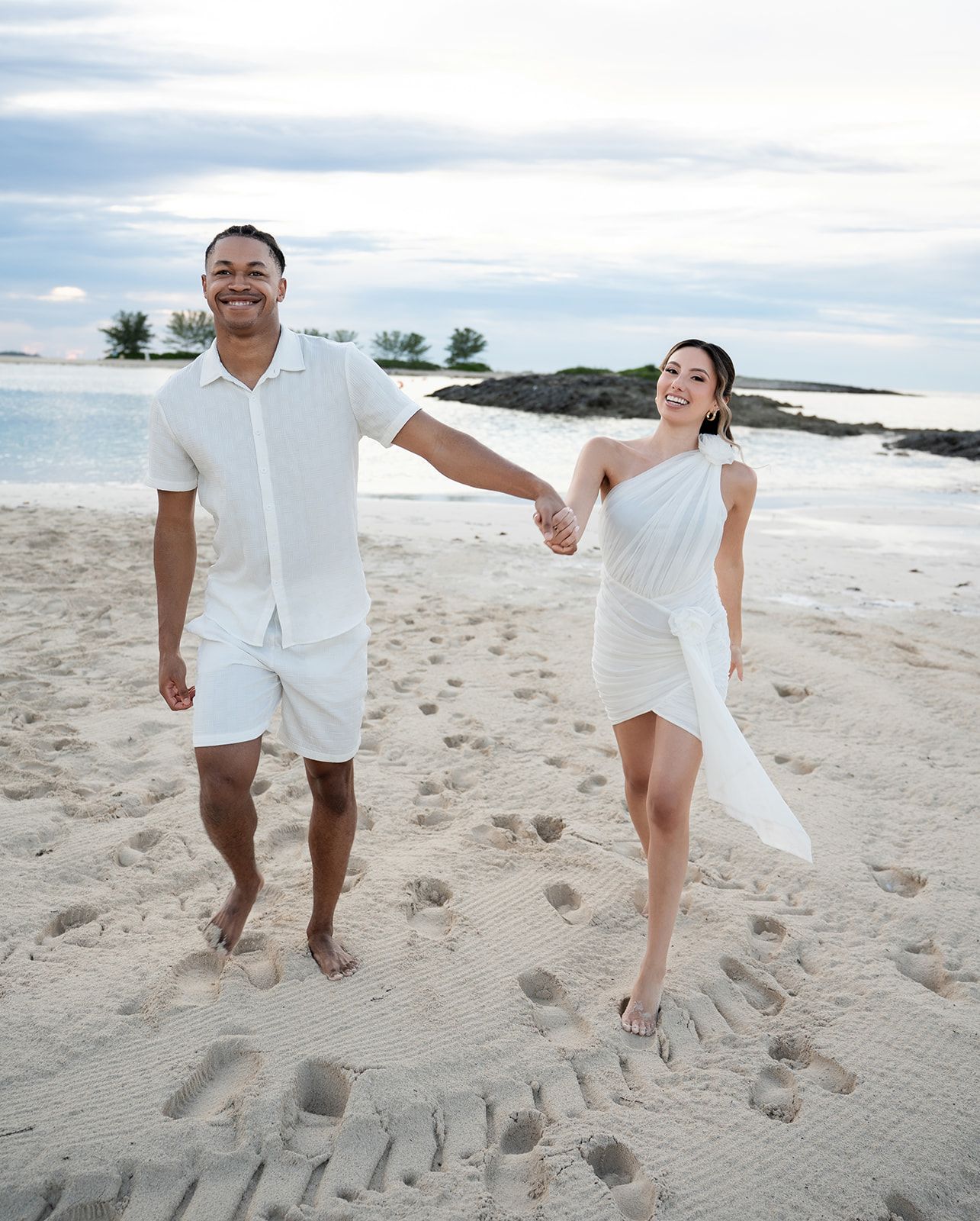 Couple dressed in white holding hands and walking barefoot on a sandy tropical beach with calm turquoise water.