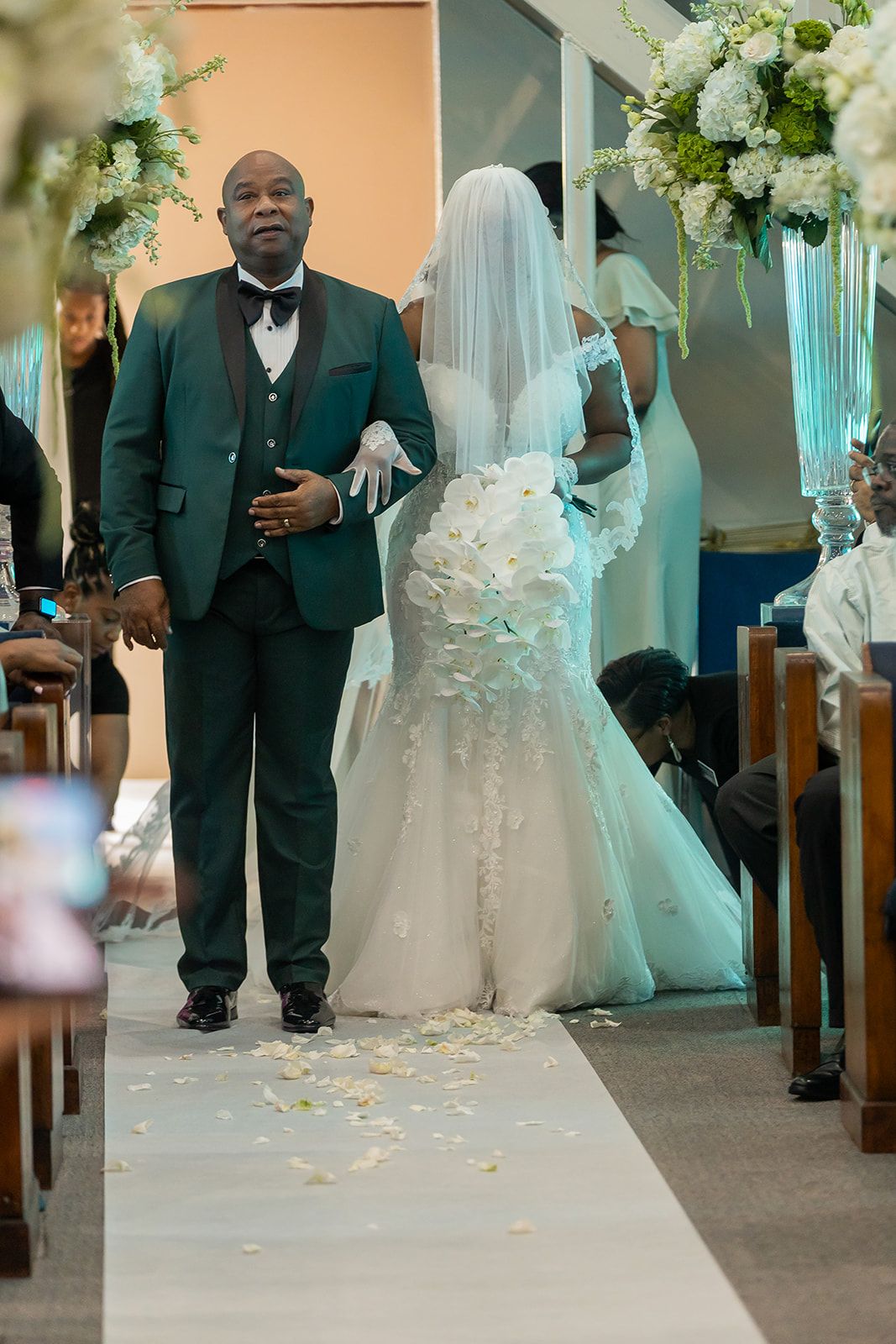 A bride in a floral white gown walks down the aisle with her escort in a green tuxedo at a church wedding.