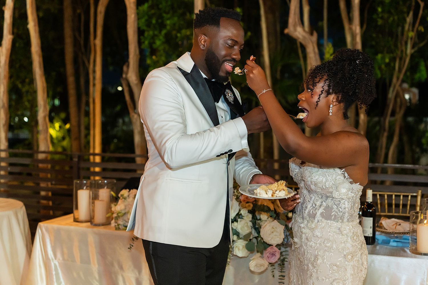 Bride and groom share a joyful cake-feeding moment at their elegant outdoor wedding reception at night.