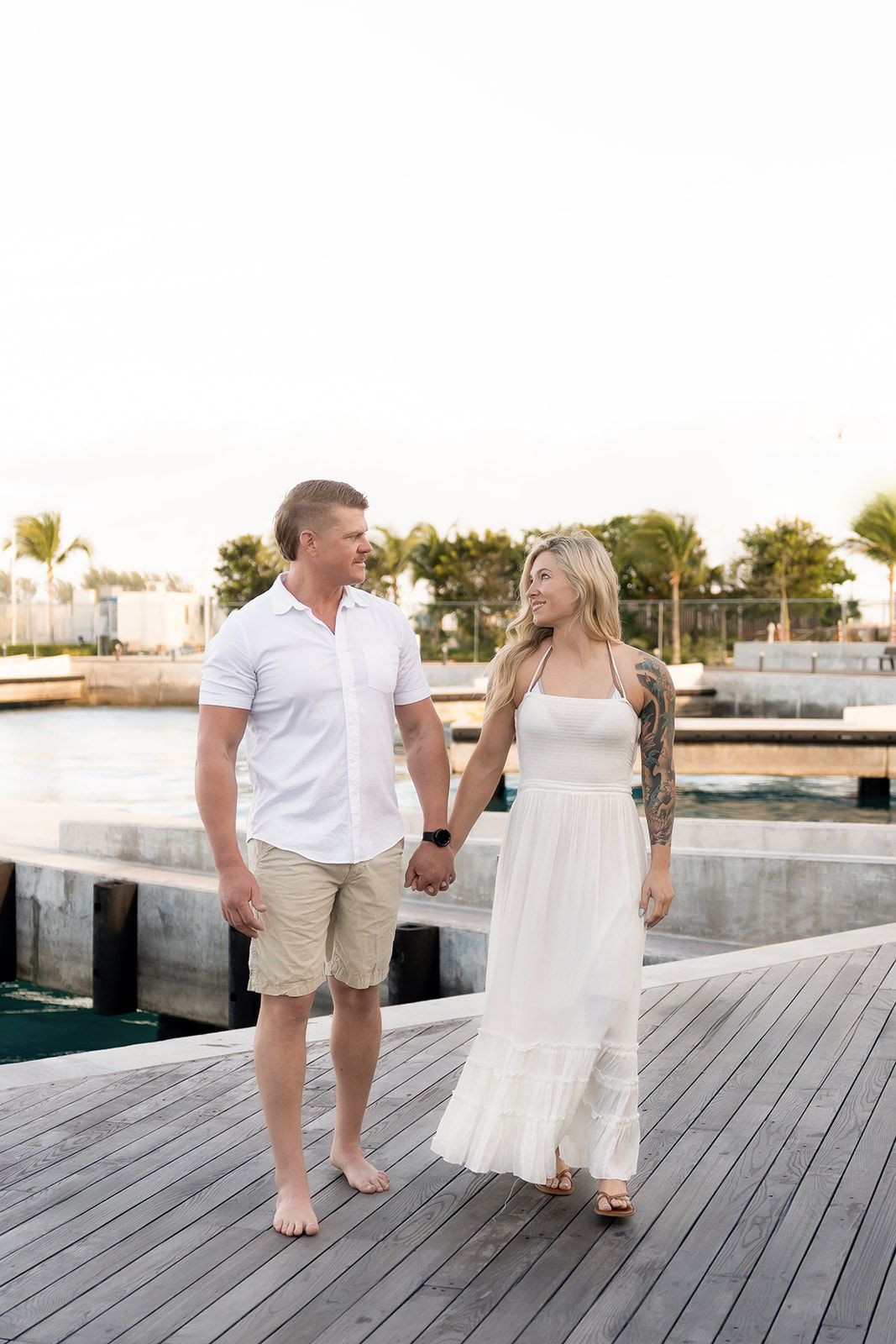 Couple holding hands on a waterfront dock, woman in white dress, man in white shirt and khaki shorts, palm trees background.