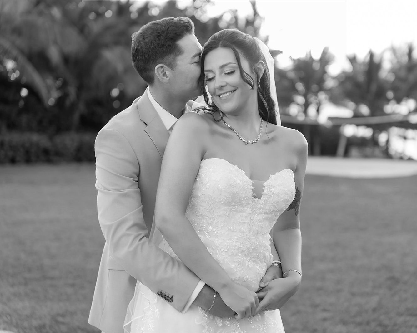 Black and white photo of a groom in a suit kissing his bride in a strapless lace wedding dress outdoors.