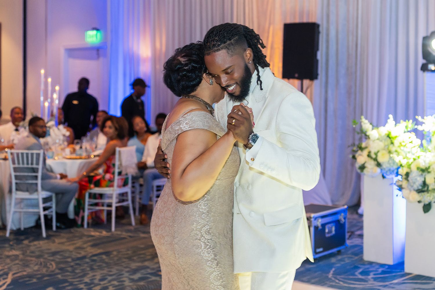 Groom and mother share a tender dance at elegant wedding reception with white floral decor.