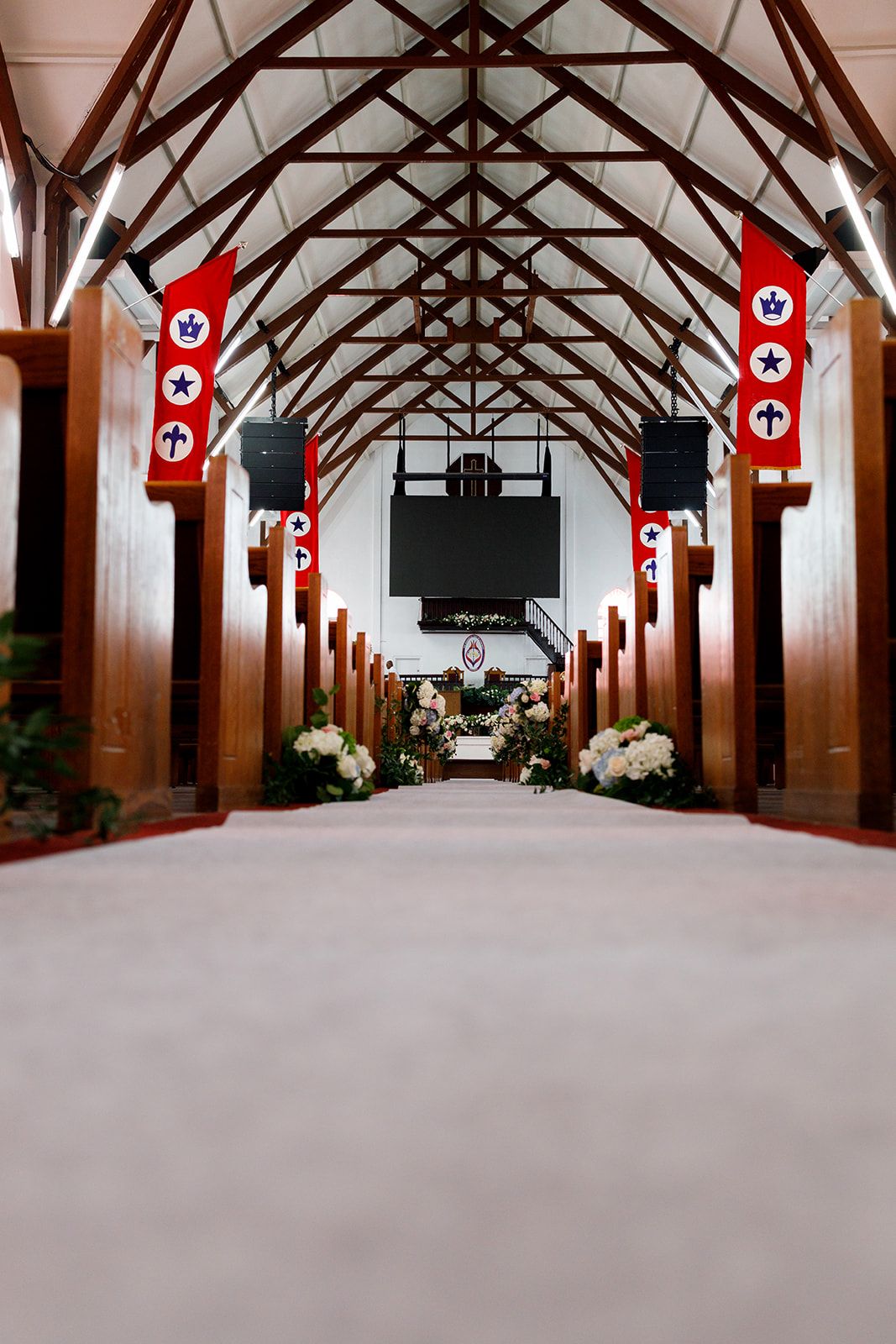 Church aisle with white carpet, wooden pews, red banners, and floral arrangements leading to altar.