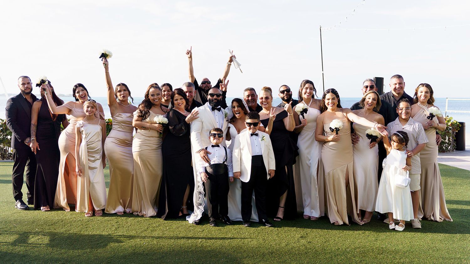 Large wedding party poses outdoors on green lawn, bridesmaids in champagne gowns, groomsmen in suits, celebrating joyfully.