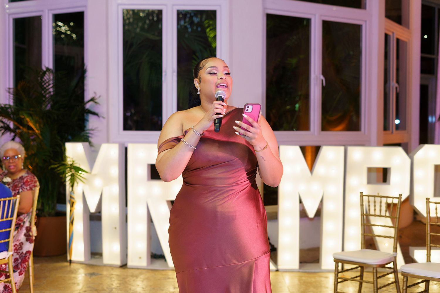 Woman in pink satin dress speaking at wedding reception with MR & MRS marquee letters in background.