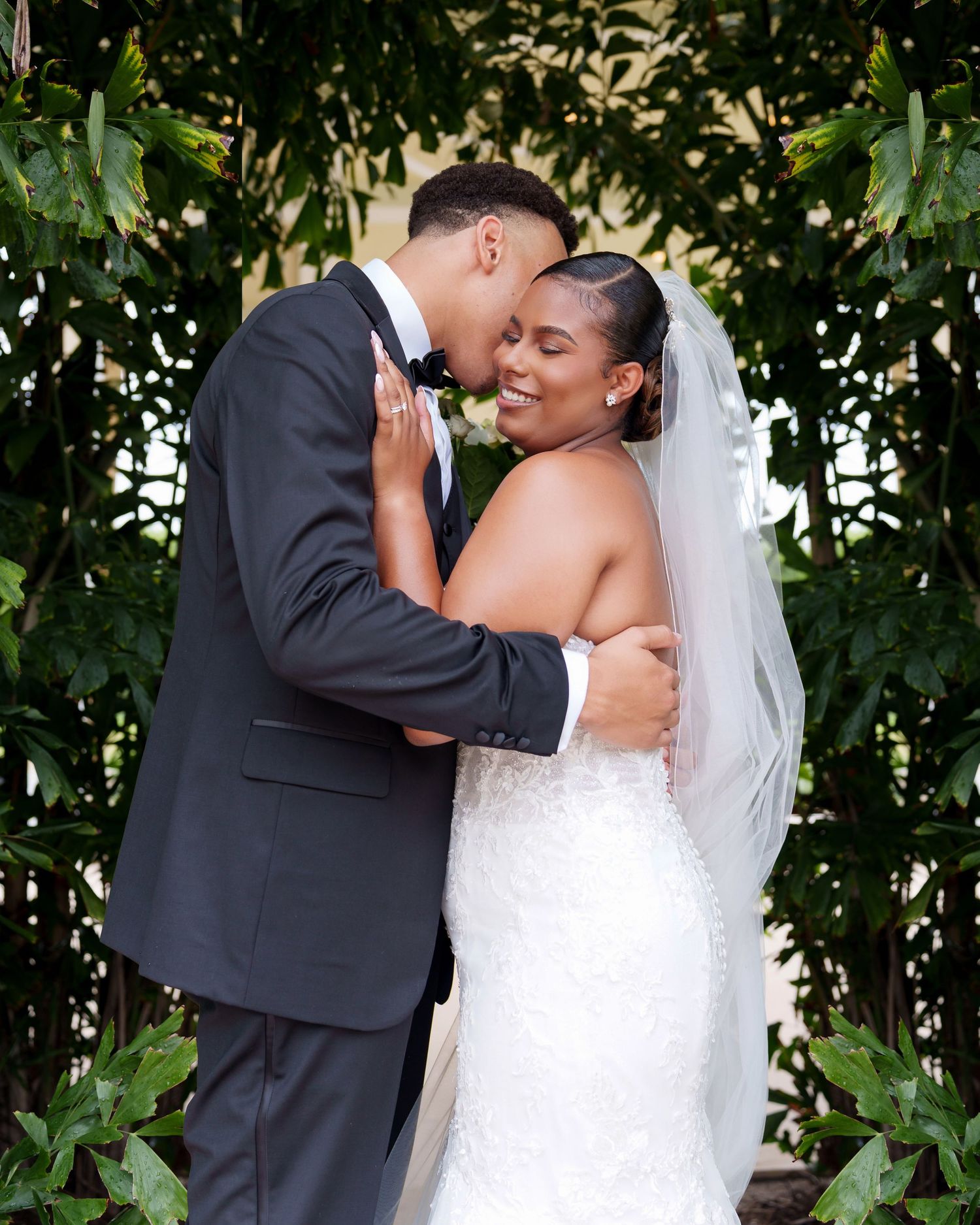 A bride and groom share a romantic embrace outdoors, surrounded by lush green foliage on their wedding day.