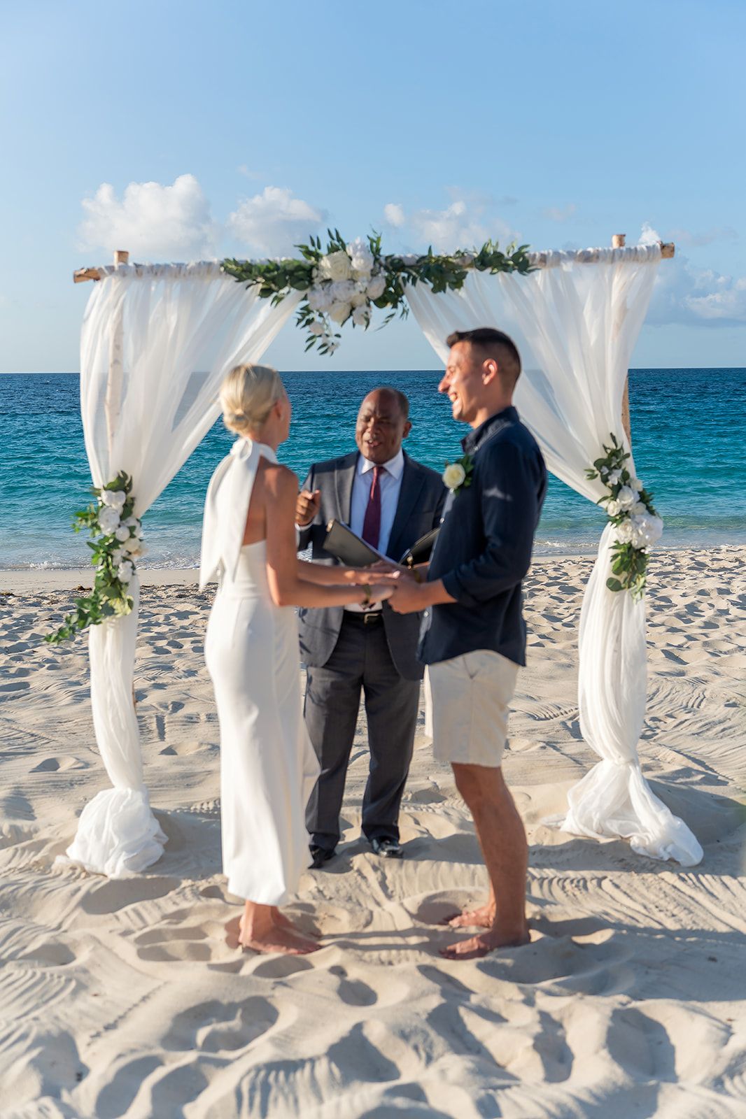 Couple exchanging vows at a beach wedding ceremony under a white floral arch with ocean backdrop.