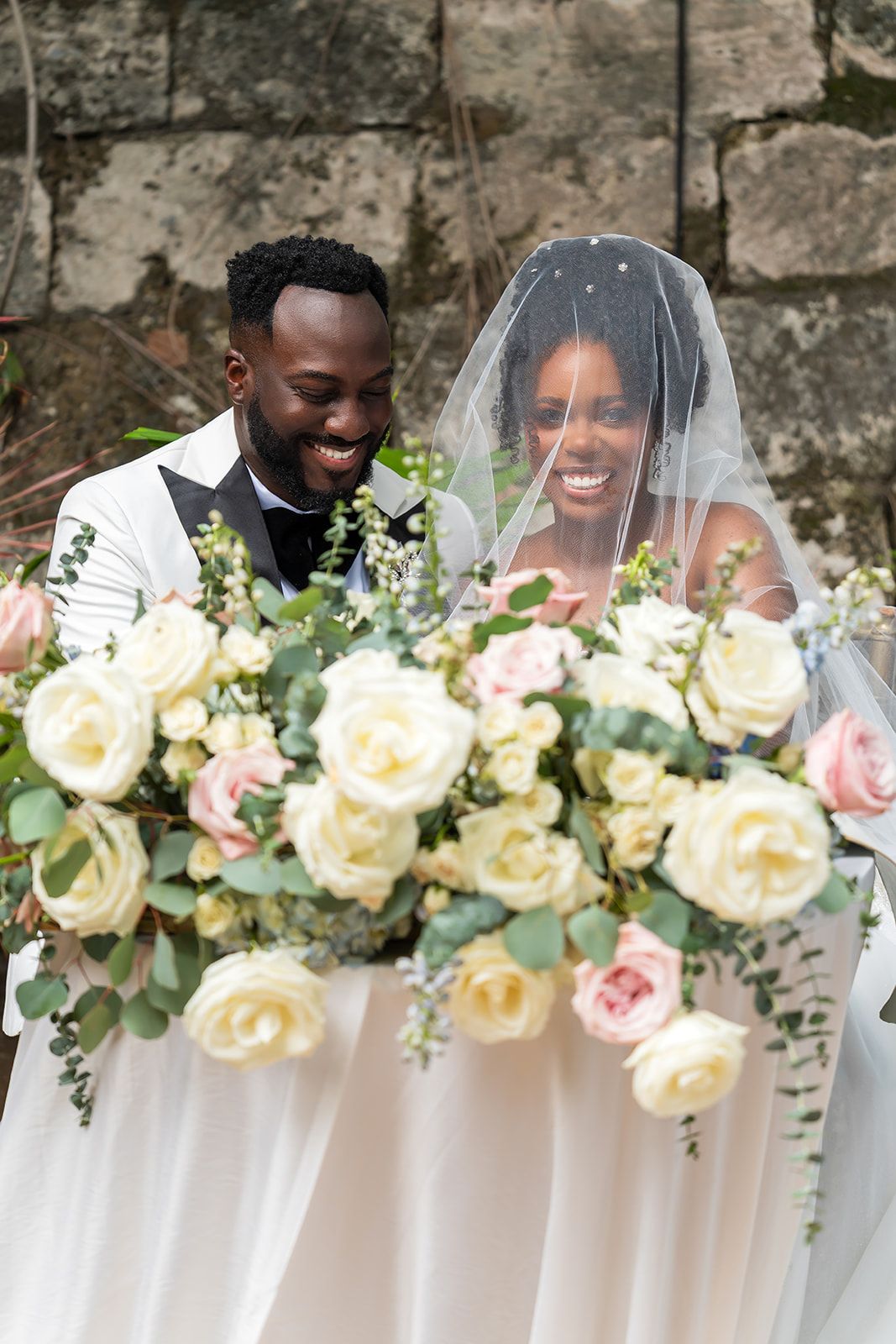 Smiling bride and groom surrounded by lush white and pink rose bouquets on their wedding day.