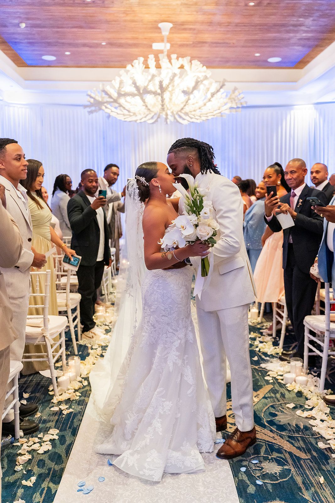 Bride and groom kissing at wedding ceremony altar with guests celebrating in elegant chandelier ballroom.