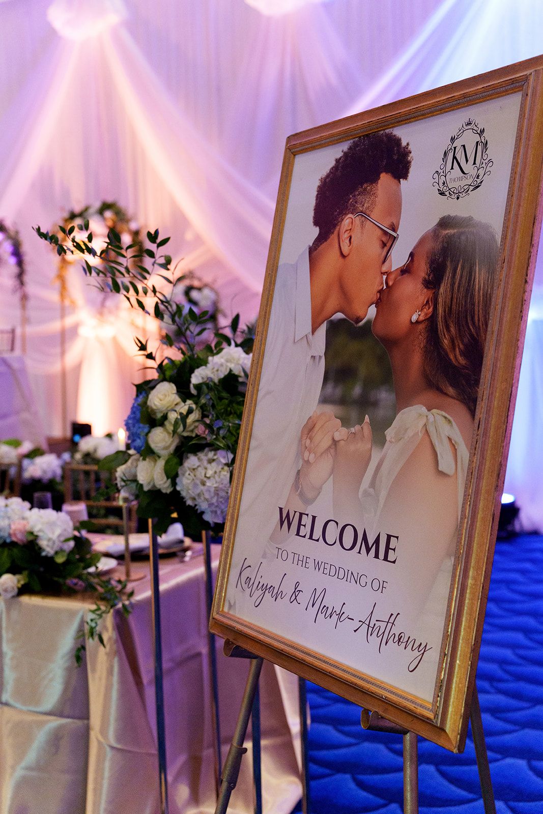Wedding welcome sign for Kaliyah and Mark-Anthony featuring couple kissing photo with floral arrangements.