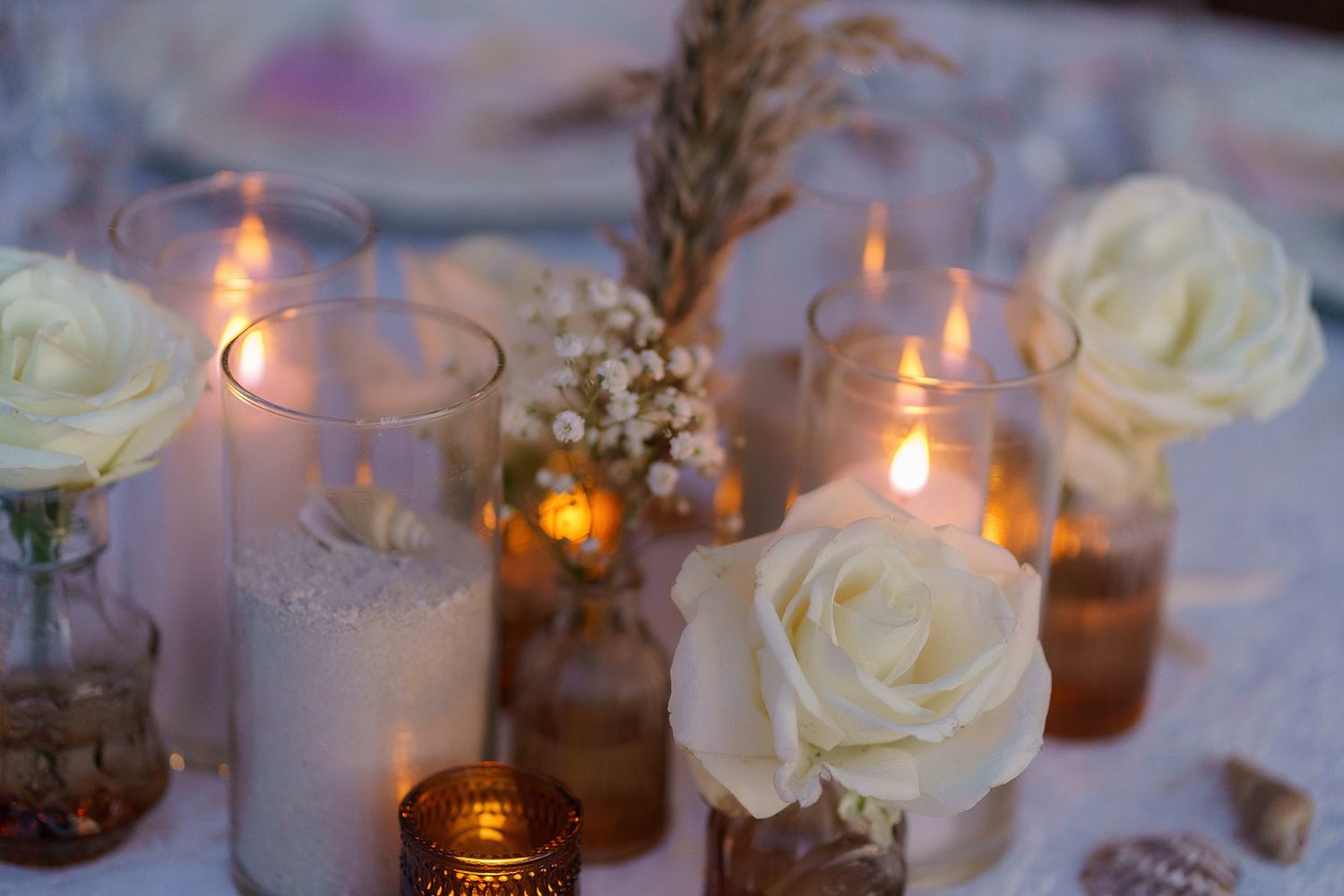 Elegant wedding table centerpiece with white roses, baby's breath, and glowing candles in glass vases.