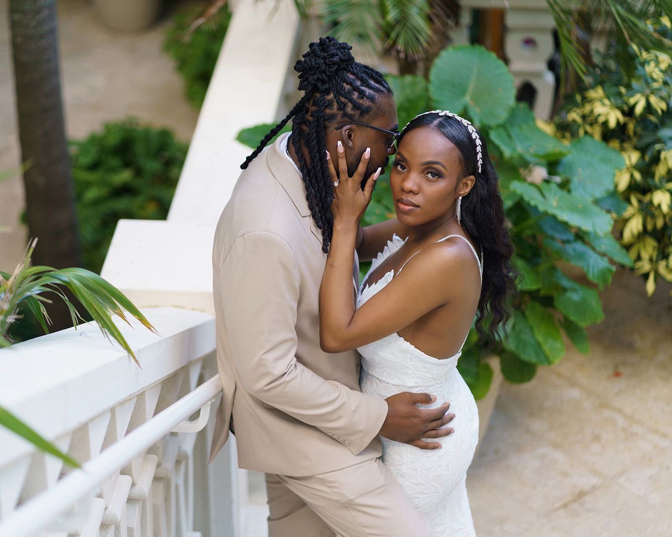 A couple in wedding attire embraces on a balcony surrounded by lush tropical greenery.