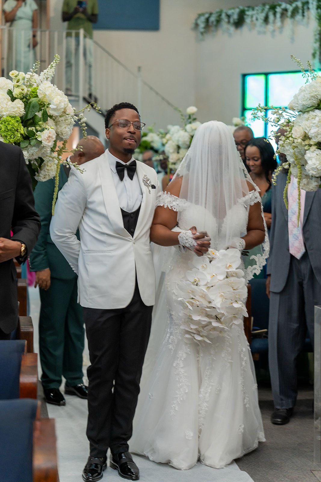 Groom in white tuxedo jacket walks bride in lace gown down church aisle surrounded by white floral arrangements.