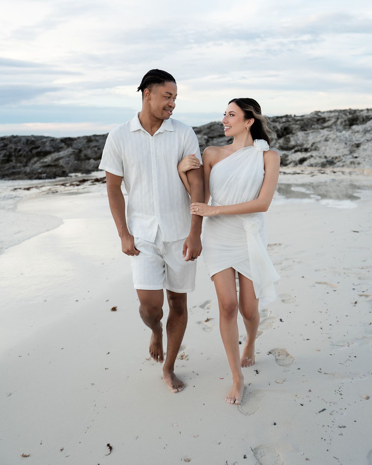 Couple in matching white outfits walking barefoot on a sandy beach, smiling at each other near rocky shores.