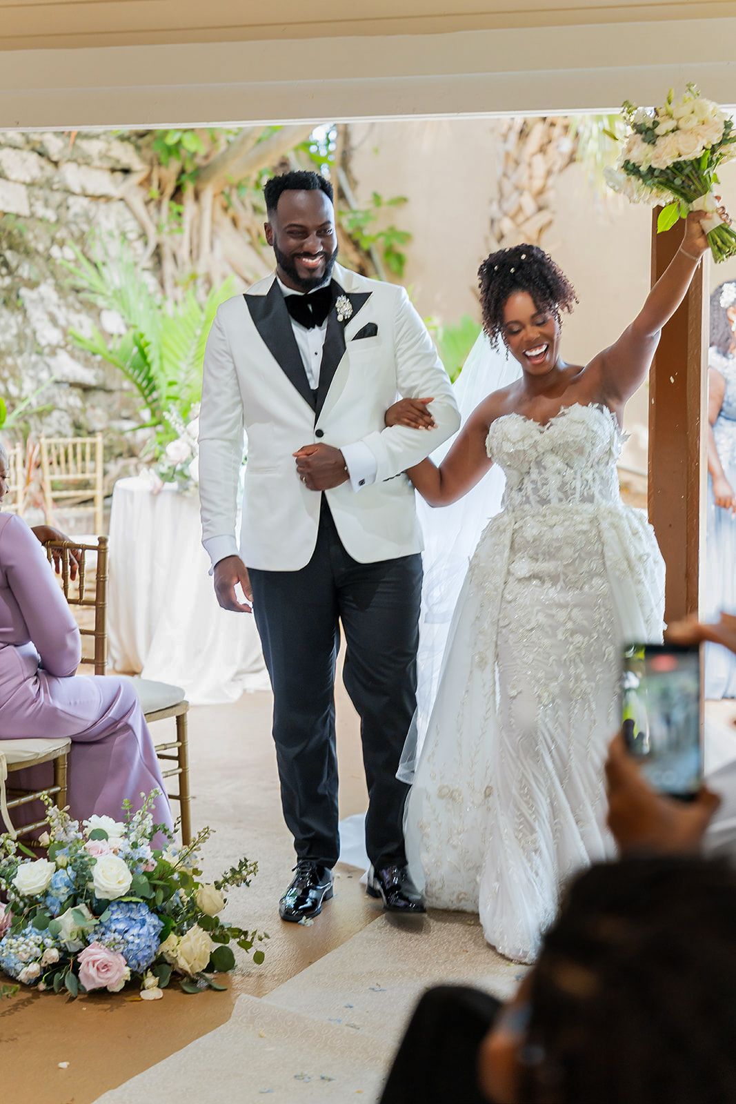 Joyful newlyweds walk down the aisle, bride in lace gown raising bouquet, groom in white tuxedo jacket.