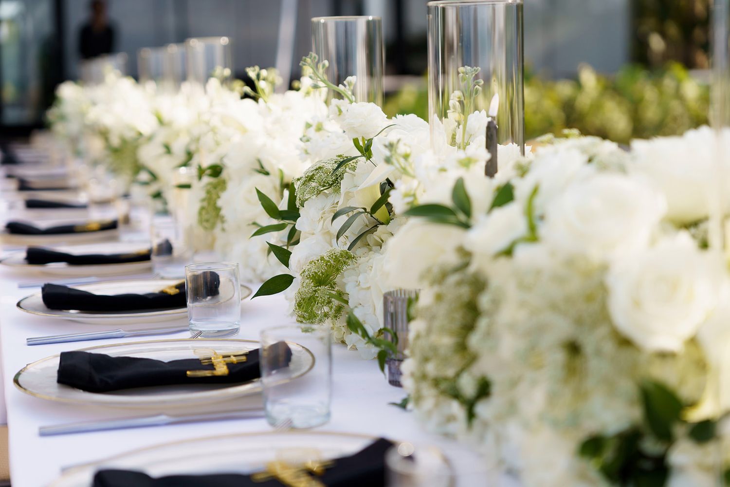 Elegant wedding reception table with white floral centerpieces, black napkins, and tall glass candle holders.