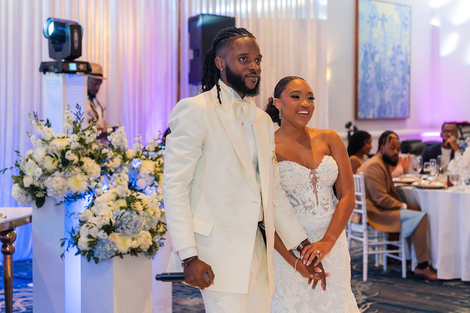 Bride and groom in elegant white attire smile together at their wedding reception with floral arrangements.