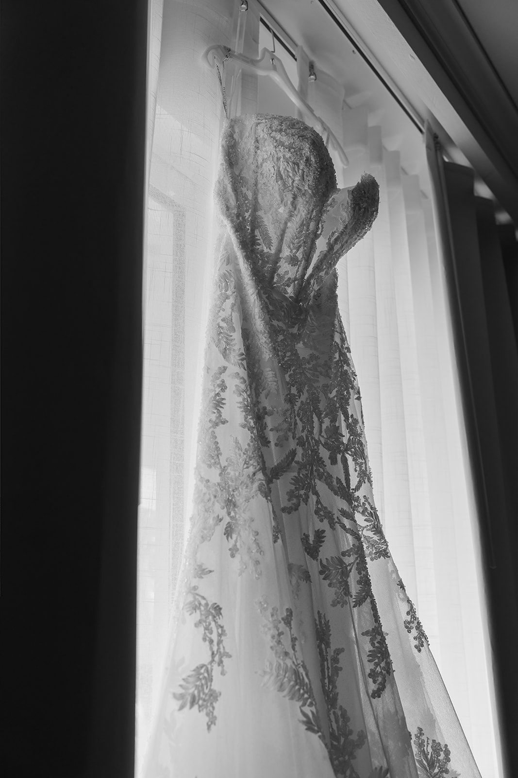 Black and white photo of an elegant lace wedding dress hanging by a sunlit window.