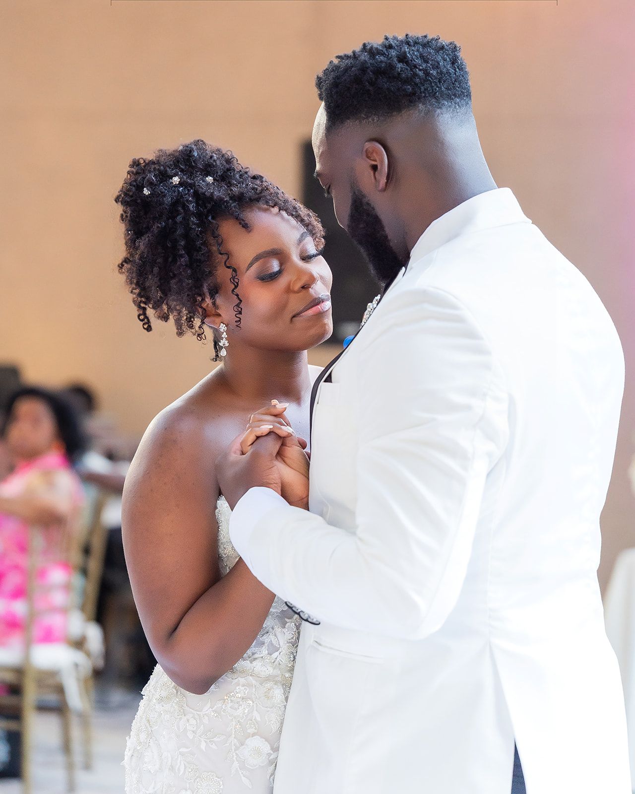 Bride and groom share intimate first dance, groom in white blazer holding bride close in elegant wedding reception.