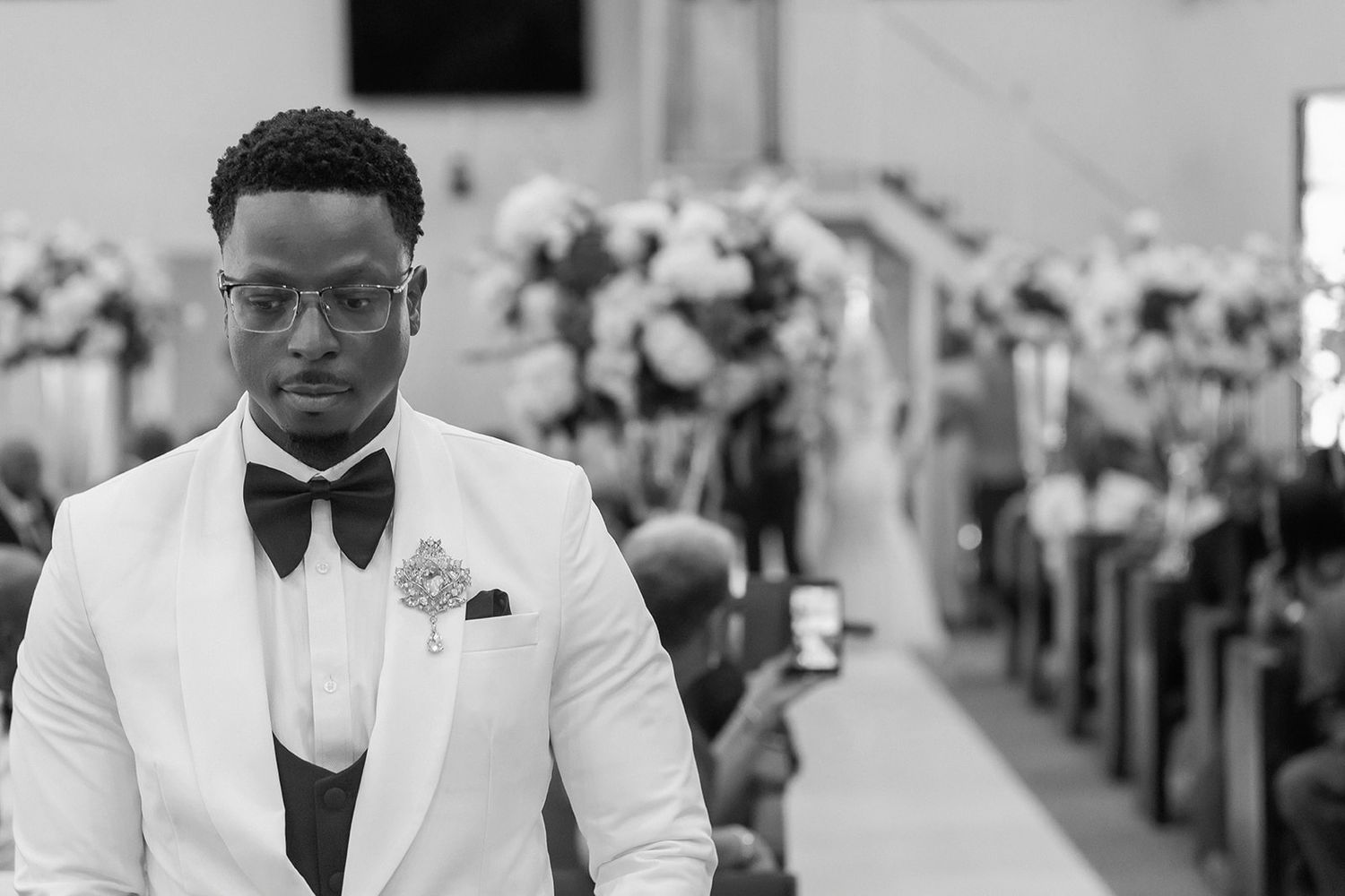 Groom in white tuxedo with bow tie stands in church aisle on wedding day, black and white photo.