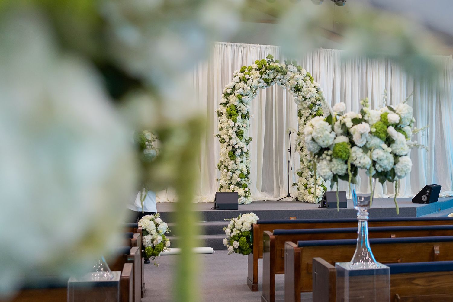 Elegant wedding ceremony setup with floral arch, white draping, and lush flower arrangements on wooden pews.