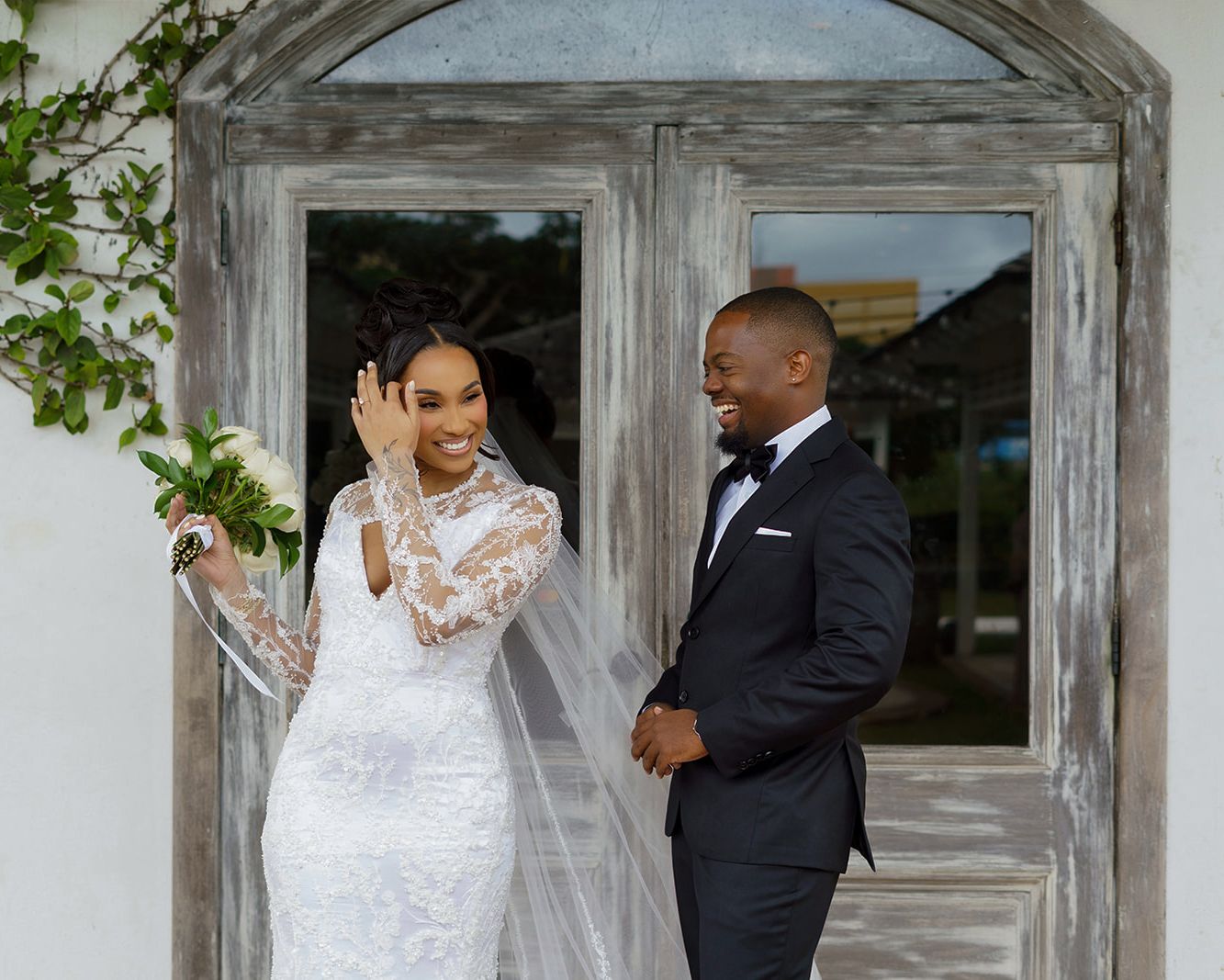 Bride in lace gown and groom in tuxedo share a joyful first look moment outside rustic wooden doors.