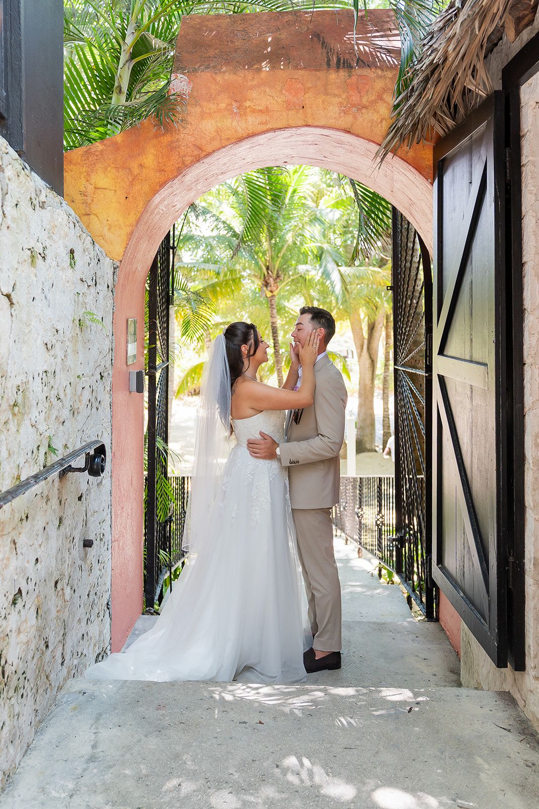 Bride and groom share a romantic kiss under a pink arched gateway surrounded by lush tropical palm trees.