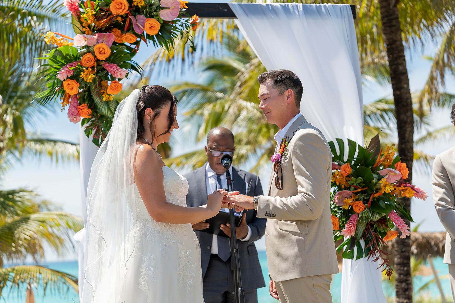 Bride and groom exchange rings under a floral arch with officiant at a tropical beach wedding.