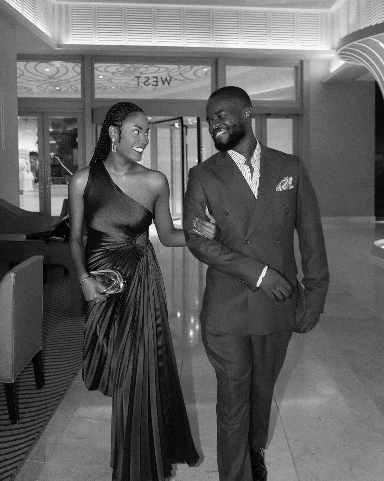 Elegant couple in black and white photo, woman in satin gown and man in suit, smiling at formal venue.