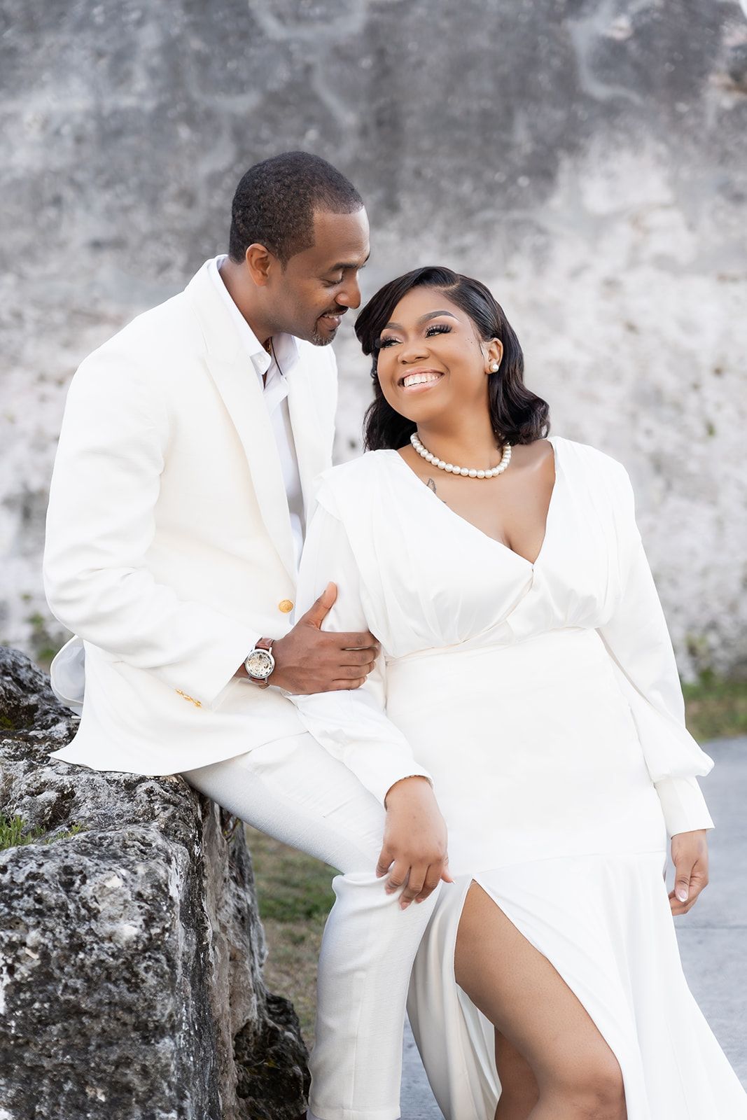 Elegant couple in white wedding attire sitting on stone ruins, smiling joyfully during romantic outdoor photo session.