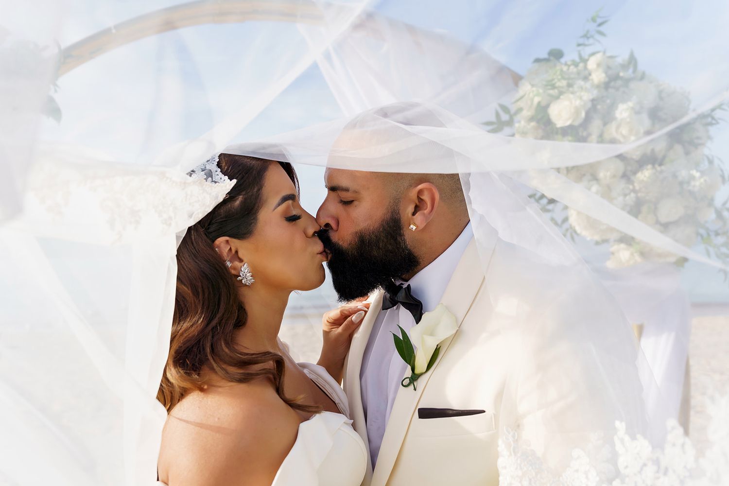 Bride and groom sharing a romantic kiss under a flowing white veil on their wedding day.