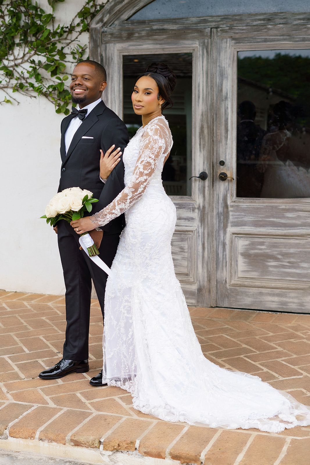 A bride in a white lace gown and groom in a black tuxedo pose together outside rustic doors on their wedding day.