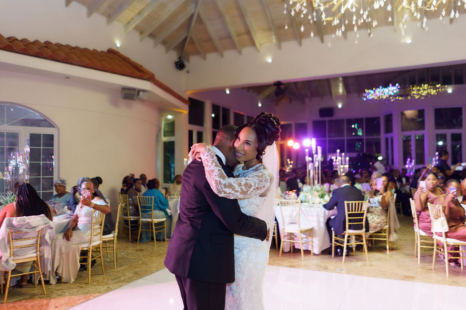 Couple sharing first dance at elegant wedding reception with guests seated at decorated tables.