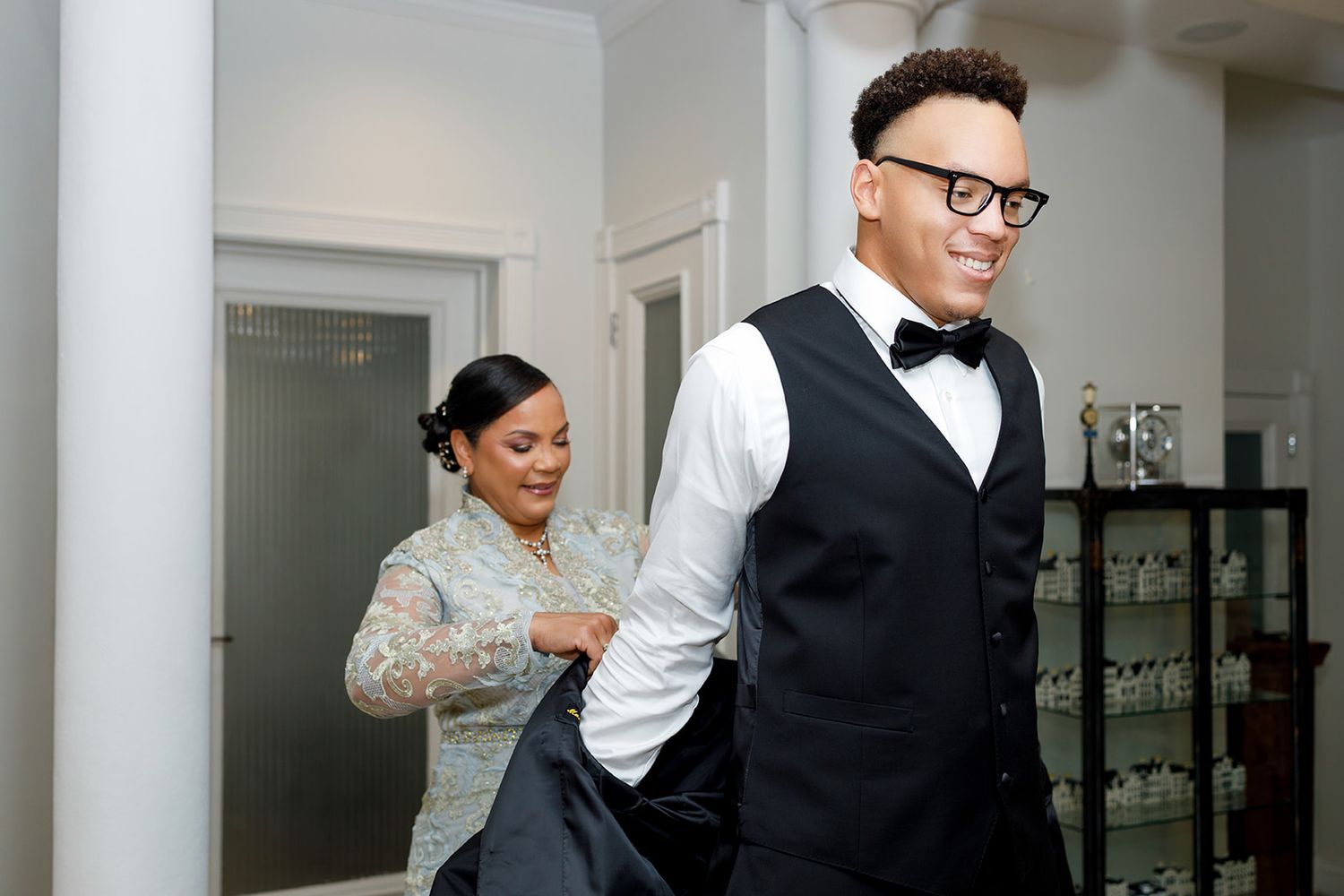 A woman helps a groom in a black tuxedo and bow tie put on his jacket indoors before a wedding.