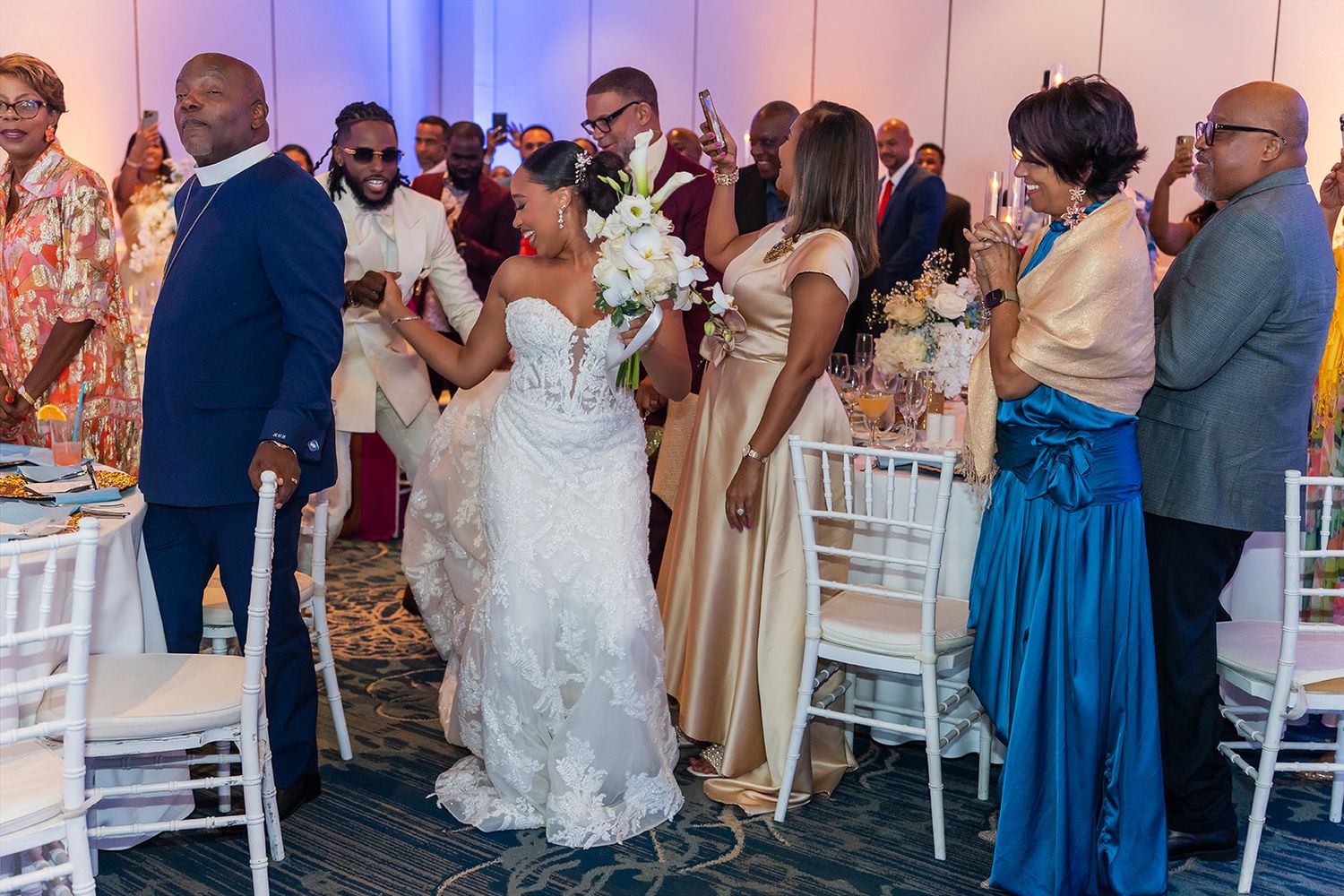 Bride in white gown holds bouquet surrounded by guests in elegant attire at a vibrant wedding reception.