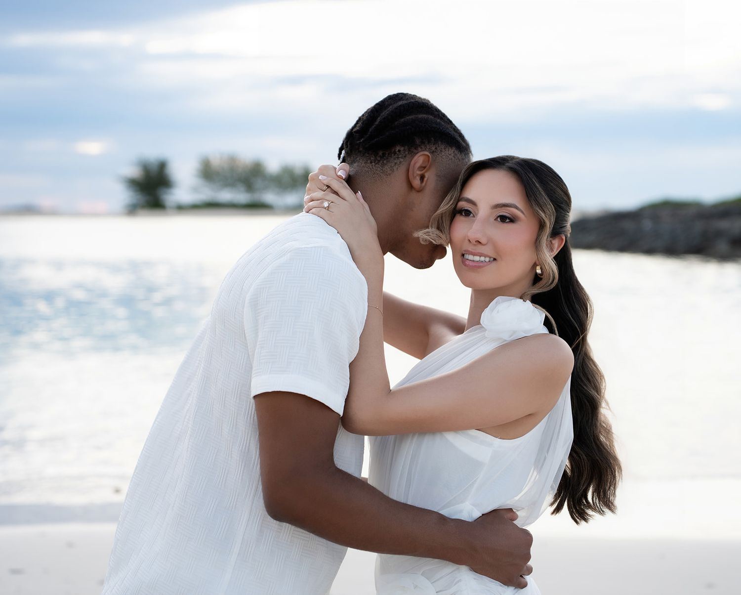 A couple embraces on a tropical beach, both wearing white outfits, with turquoise water and palm trees in the background.