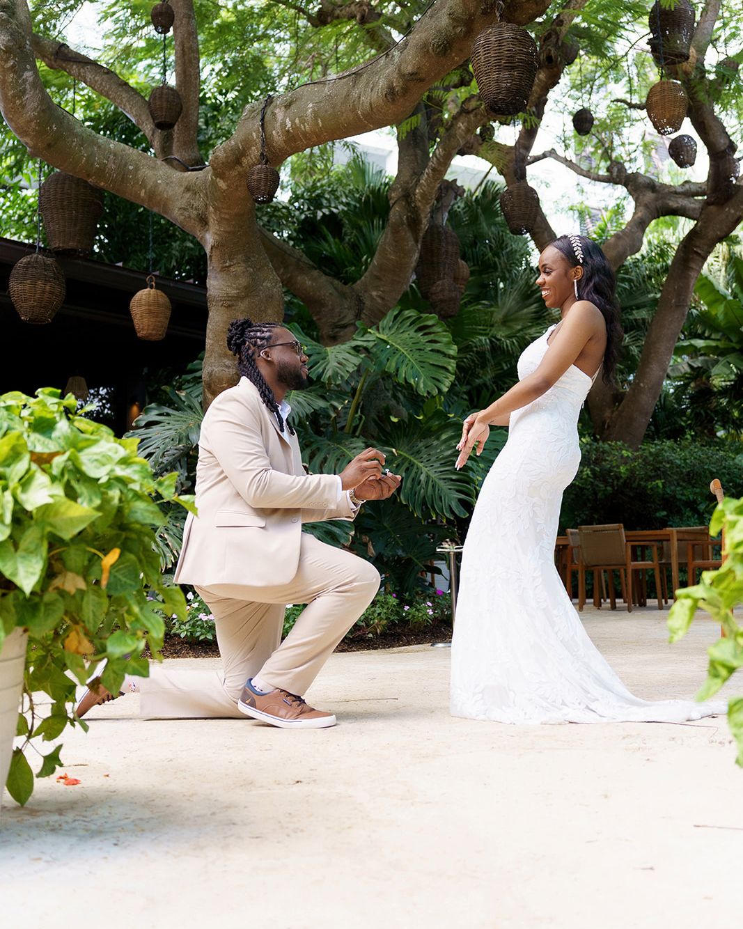 A groom kneels before his bride in a white gown during an outdoor wedding proposal under lush tropical trees.