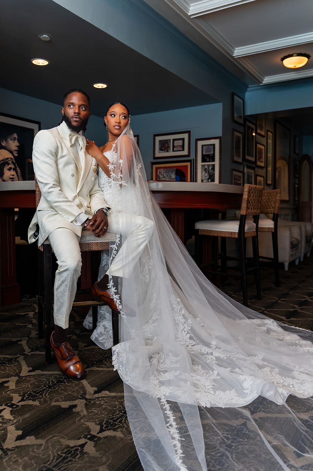 Bride and groom in white wedding attire pose elegantly with a long lace veil trailing on the floor.
