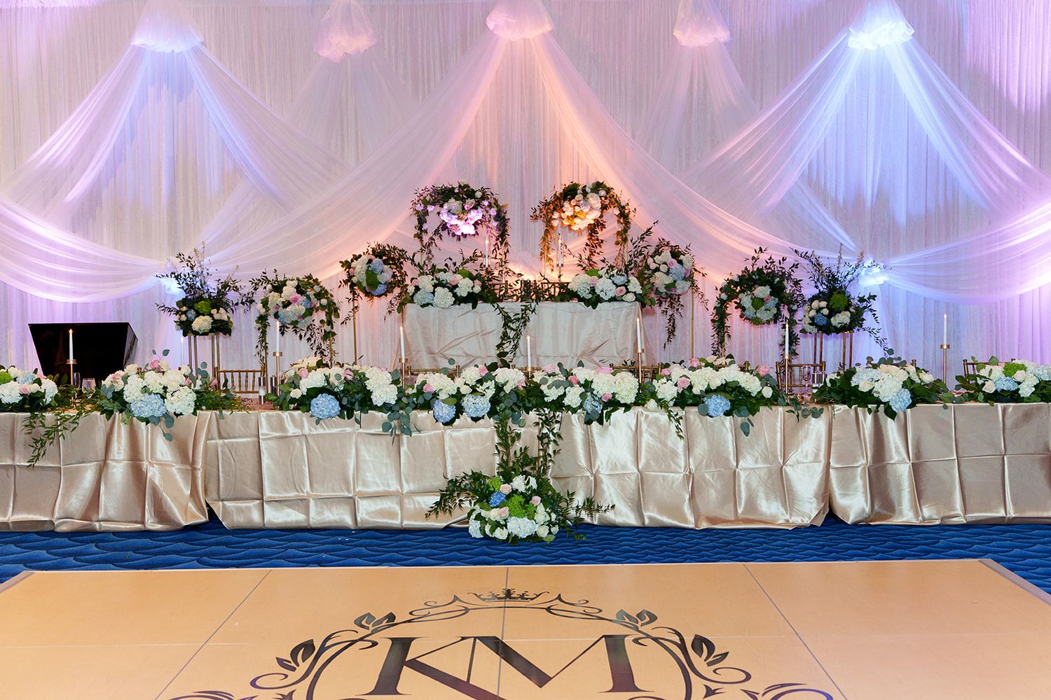 Elegant wedding head table with white draping, floral centerpieces, and KM monogram on dance floor.