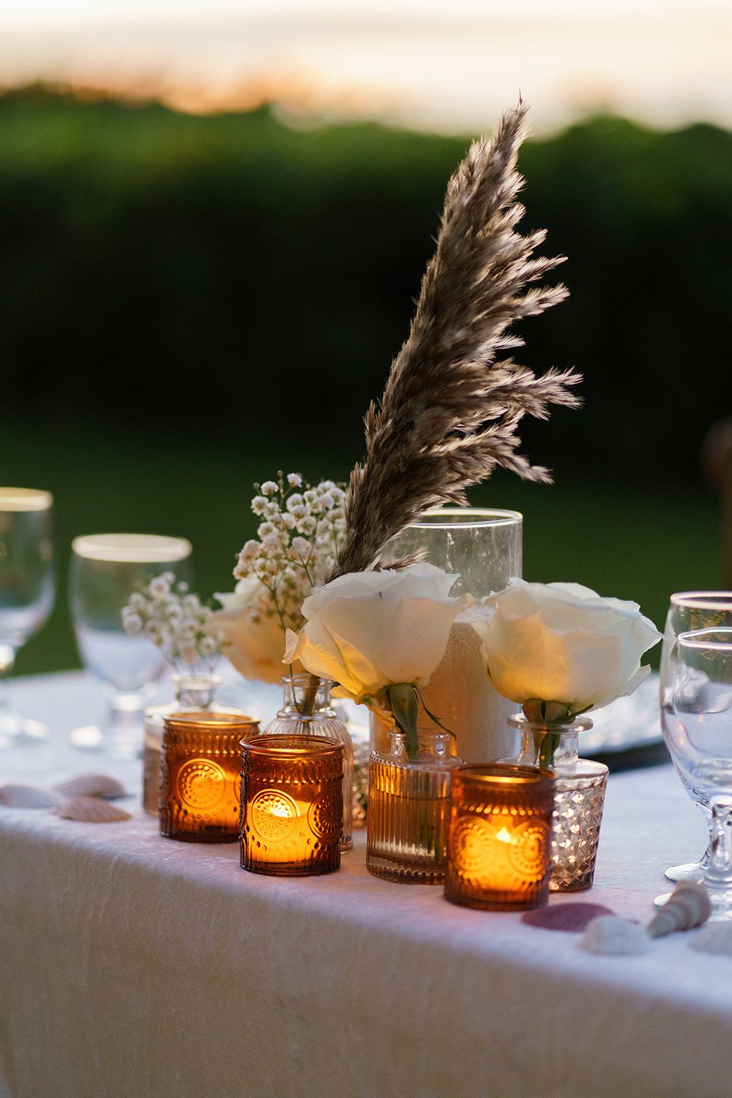 Elegant wedding table centerpiece with white roses, pampas grass, amber votive candles, and crystal glassware at dusk.