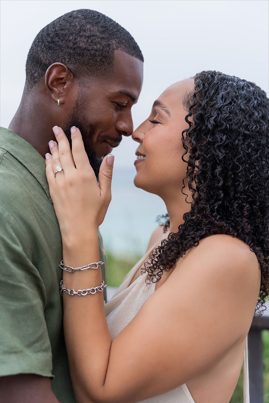 A couple shares a romantic nose-to-nose moment, woman wearing a ring and bracelet, smiling tenderly outdoors.