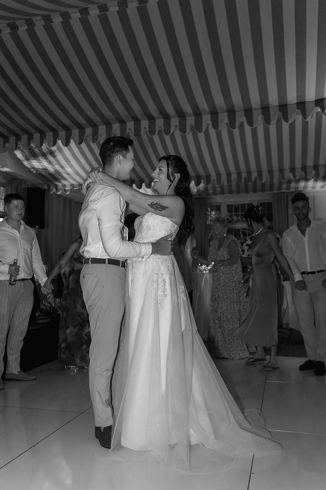 Black and white photo of bride and groom sharing their first dance at a wedding reception under a striped tent.