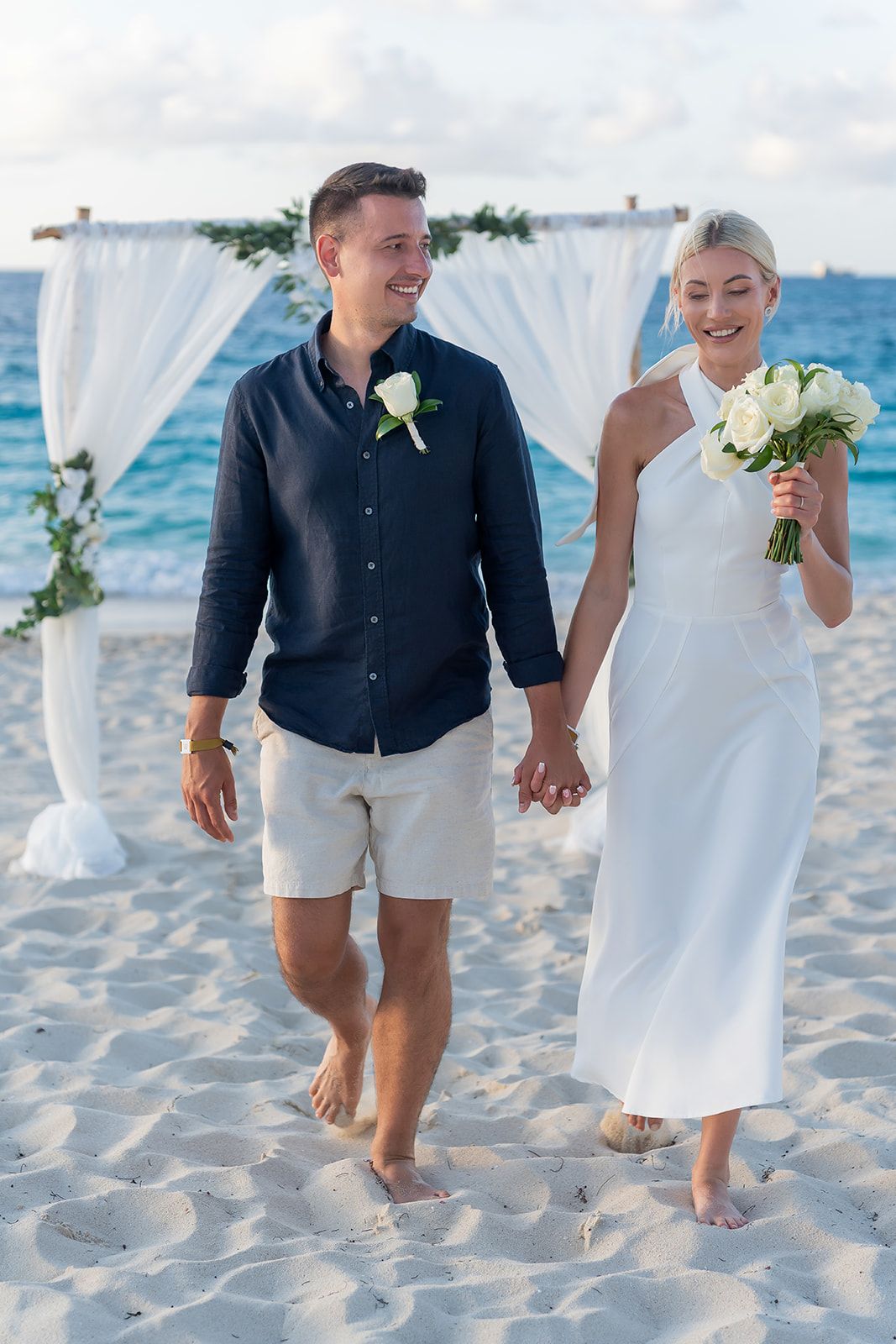 Happy newlywed couple holding hands on a sandy beach, bride in white dress with bouquet, groom in navy shirt with boutonniere.