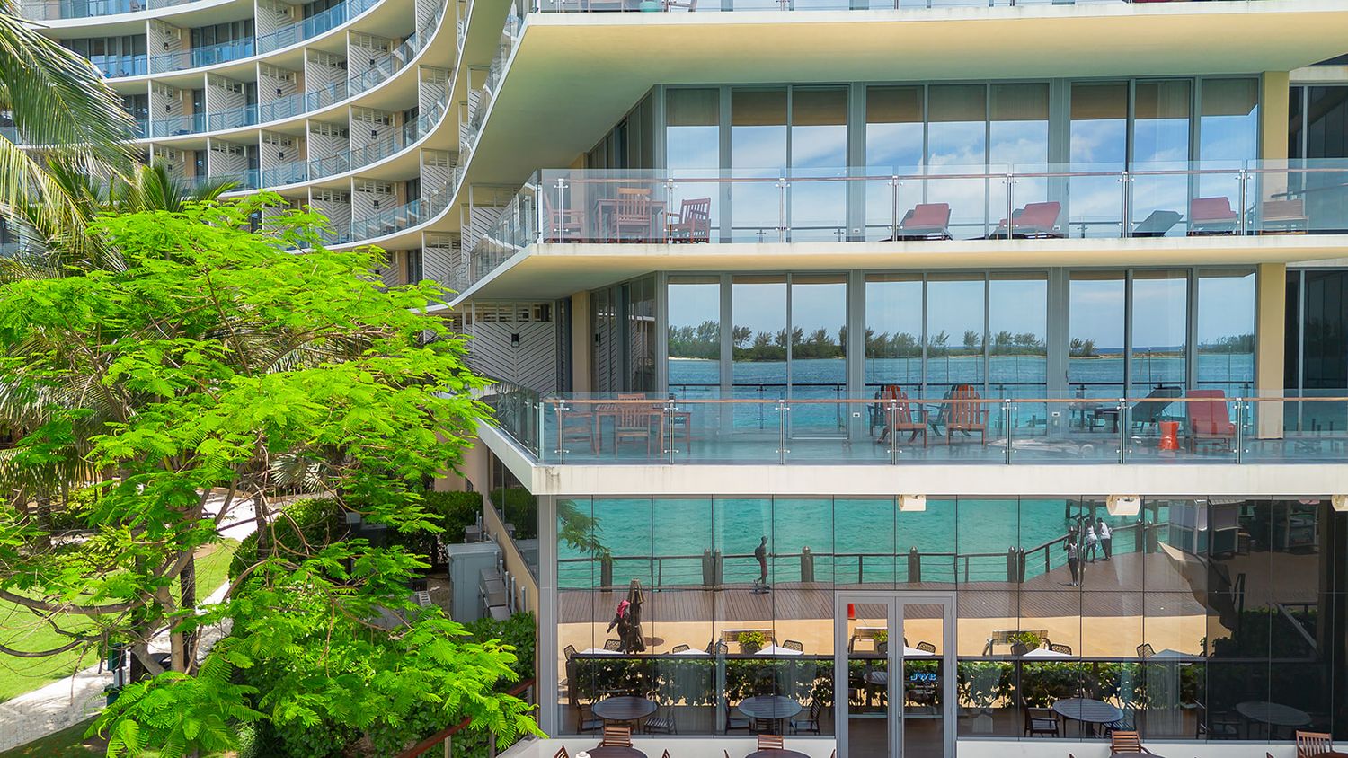Modern waterfront building with curved architecture, glass balconies, and lush palm trees in a tropical setting.